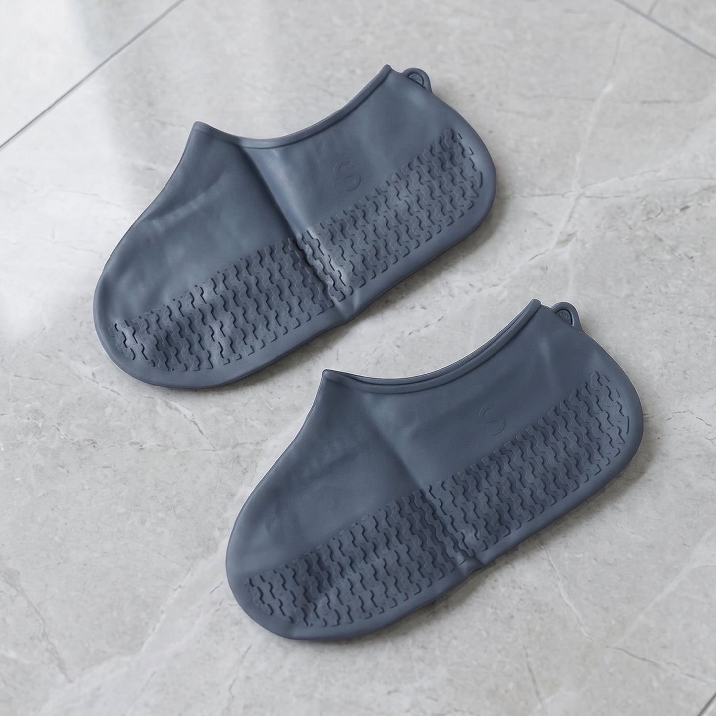 A pair of dark gray silicone shoe covers with textured soles is presented on a light gray marble-patterned floor.